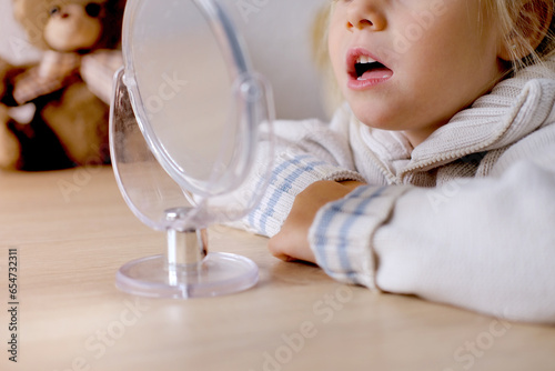 small child front mirror, blonde girl of 4 years old engaged with speech therapist, gymnastics for tongue, defect, speech disorder with frequent repetition sounds, syllables, spasms muscles apparatus photo