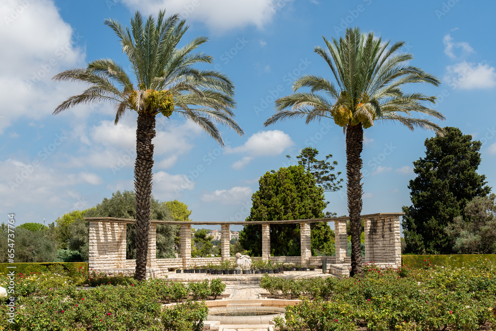 The rose garden of Park Ramat Hanadiv with a brick structure for sitting, sundial sculpture, palm trees and fountain. 
