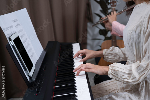 Woman in white dress press key on , tablet and key note put on  holder, and her friend playing flute beside with blurred guitar and home. Playing music together at home for fun and relaxation.