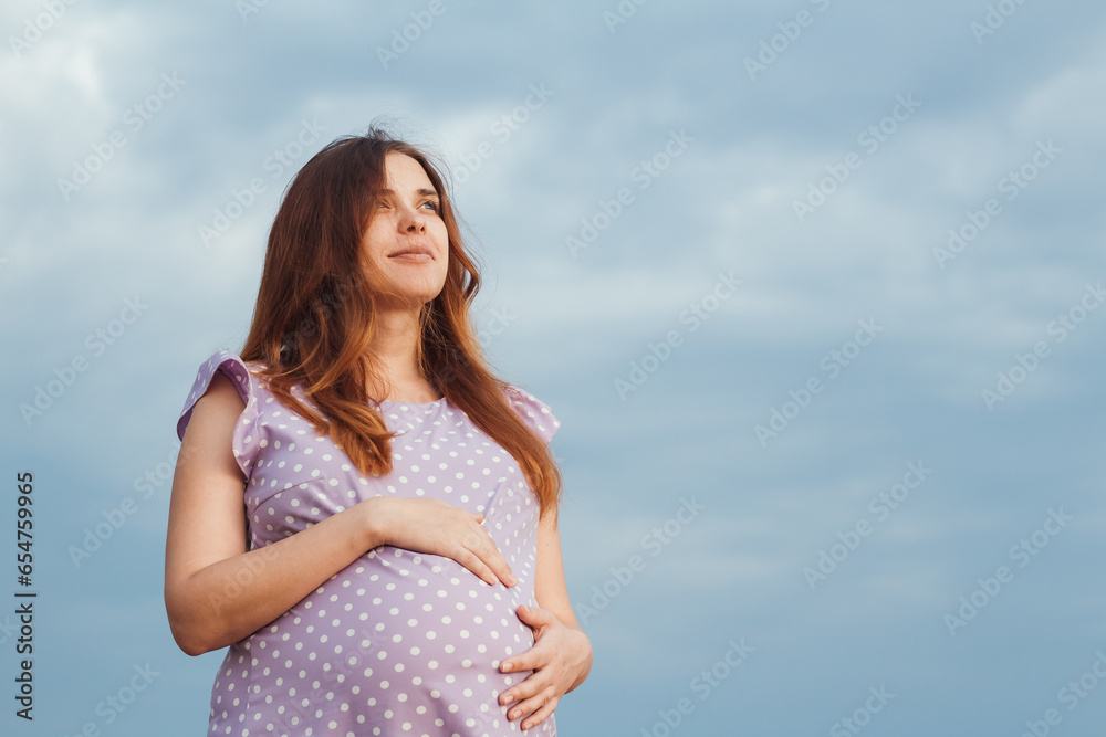 beautiful red-haired pregnant woman in summer dress stands against background of cloudy sky, girl relaxing on nature, bottom view beautiful red-haired pregnant woman in summer dress stands against background of cloudy sky, girl relaxing on nature, bottom view