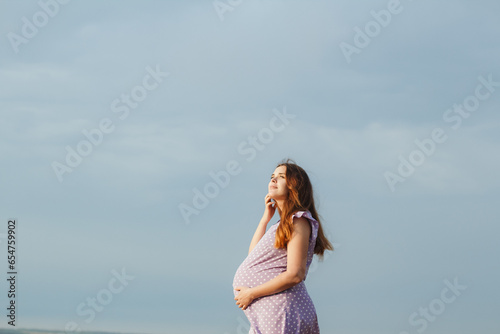 beautiful red-haired pregnant woman in summer dress stands against background of cloudy sky, girl relaxing on nature, bottom view