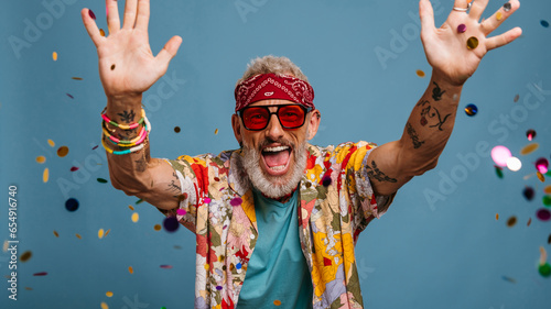 Joyful senior man in funky shirt and bandana throwing colorful confetti on blue background © gstockstudio