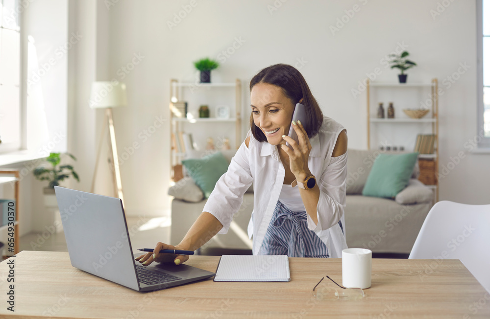 Friendly senior female entrepreneur having a business conversation on a mobile phone in her home office. Active woman makes notes and checks information using laptop. Remote business concept. Friendly senior female entrepreneur having a business conversation on a mobile phone in her home office. Active woman makes notes and checks information using laptop. Remote business concept.