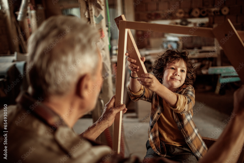 Senior male carpenter showing his grandson a wood frame in a carpentry shop Senior male carpenter showing his grandson a wood frame in a carpentry shop