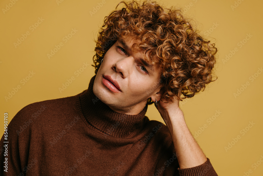 Handsome young curly man in sweater holding hand ion hair against yellow background Handsome young curly man in sweater holding hand ion hair against yellow background