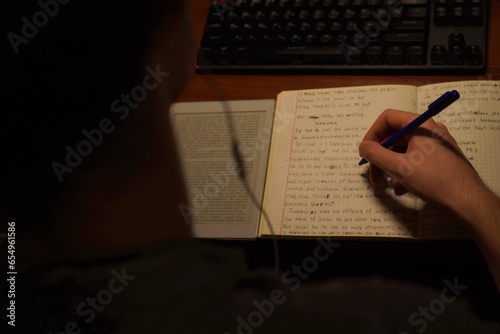 a young student performs an English language assignment in writing in a notebook using a pen and an e-book while sitting at a table in front of a computer