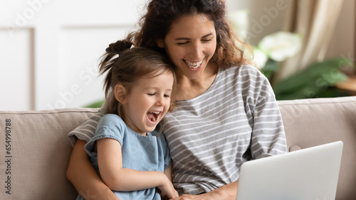 Overjoyed young mother and funny little daughter sit rest on sofa at home laugh watch funny video on laptop, happy millennial mom and small girl child relax on couch have fun using computer together