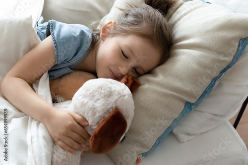 Cute little preschooler girl lying in cozy bed hugging taking nap with favorite plush toy, small kid sleep relax in comfortable bedroom cuddling with fluffy teddy bear at home, relaxation concept