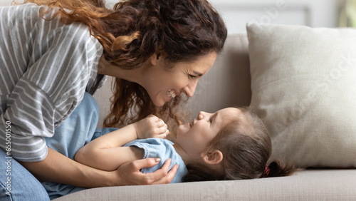 Happy young mother sit rest on sofa in living room playing with little preschooler daughter, smiling millennial mom or nanny feel playful overjoyed engaged in funny game with small girl child at home photo