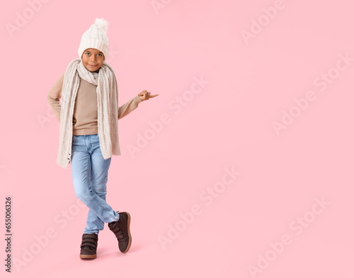 Cute African-American boy in warm winter clothes pointing at something on pink background