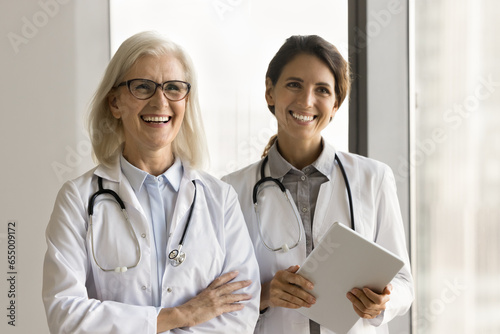 Two happy confident senior and younger doctor women in white uniform coats posing together indoors, looking away with toothy smiles, enjoying professional meeting. Portrait of medical colleagues