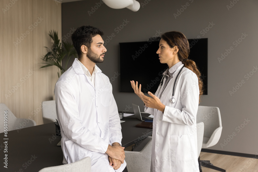 Fototapeta premium Two serious male and female doctor colleagues in white coats talking in meeting room, discussing medical practice, clinic work process, treatments cases, physician occupation, job