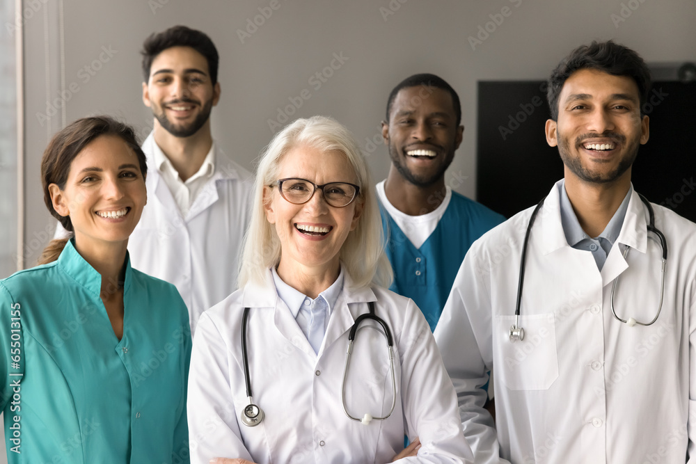 Happy mature older clinic mentor woman and multiethnic team of interns looking at camera with toothy smiles, posing for shooting. Group of positive doctors in white coats and uniforms portrait Happy mature older clinic mentor woman and multiethnic team of interns looking at camera with toothy smiles, posing for shooting. Group of positive doctors in white coats and uniforms portrait