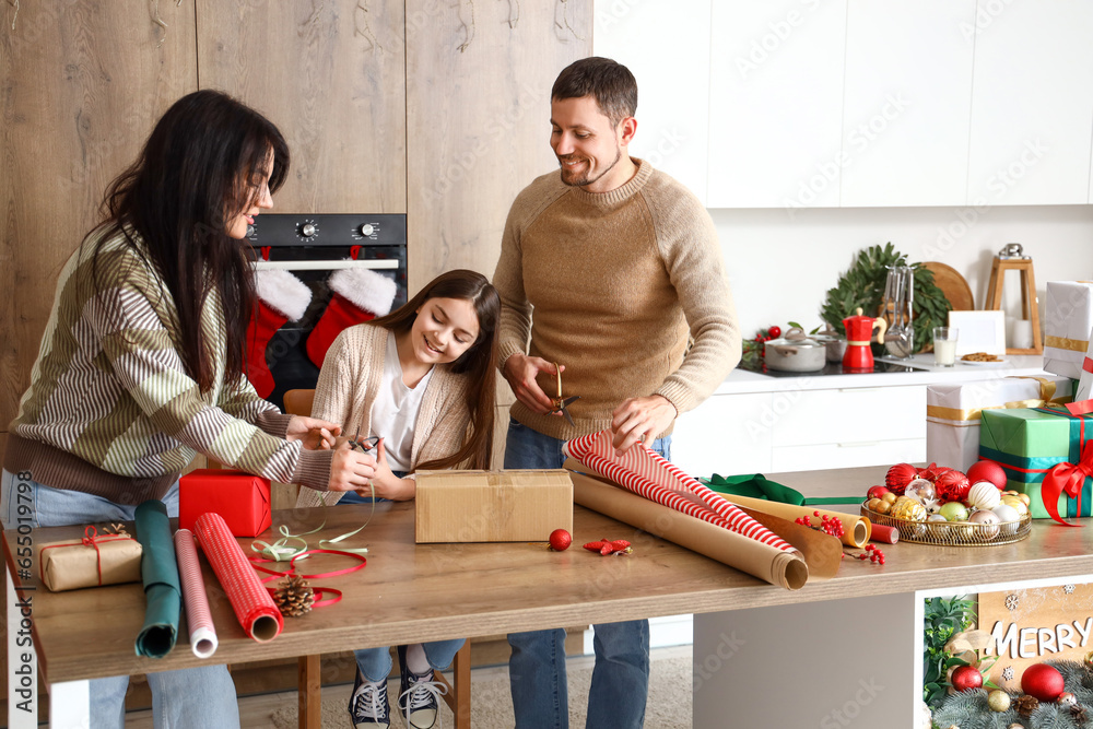 Little girl with her parents wrapping Christmas gift in kitchen - obrazy, fototapety, plakaty 