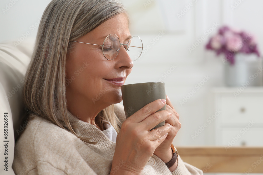 Beautiful senior woman drinking tea on sofa at home, space for text