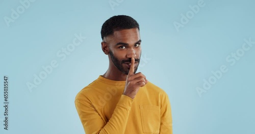 Secret, face and man with finger on lips in studio for quiet, privacy or hush news on blue background. Whisper, drama and portrait of guy model with confidential hand emoji for gossip or announcement photo