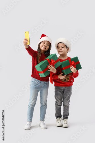 Cute little children with Christmas gifts taking selfie on light background