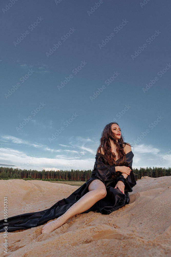 A brunette in a black dress with a long train made of silk fabric sits on a sandy dune in the wind. Landscape of sand dunes and blue sky with clouds.