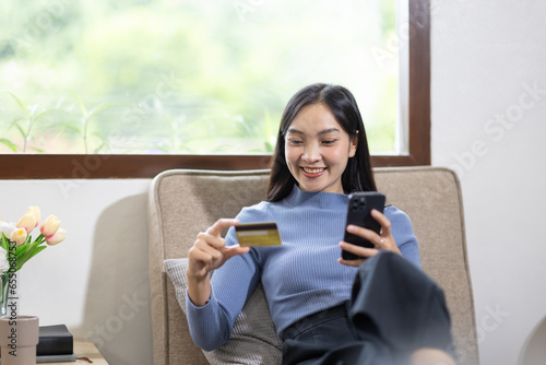 Young asian woman holding credit card and shopping or online payment by using nusing phone laptop computer. Businesswoman at home. Online shopping, e-commerce, internet banking, spending money.