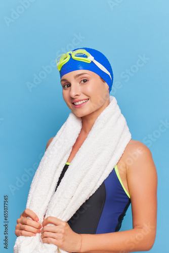 Pretty woman in swimsuit and bathing cap stands half-turned isolated on blue background holding towel around her neck with her hands, professional sport concept, copy space