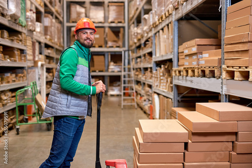 pleasant warehouse employee pushing manual pallet jack and working in factory storage room, warehouse worker transporting cardboard goods delivery. logistics and distribution concept © alfa27