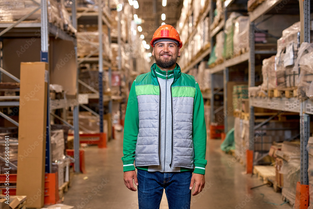 Portrait of happy smiling young warehouse worker posing posing near shelves with goods. Feeling satisfied doing job good. Young employee in working uniform and safety helmet, look at camera Portrait of happy smiling young warehouse worker posing posing near shelves with goods. Feeling satisfied doing job good. Young employee in working uniform and safety helmet, look at camera