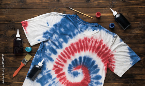 A white T-shirt painted in red and blue tie dye style and materials for coloring on a wooden background. The concept of self-dyeing clothes.