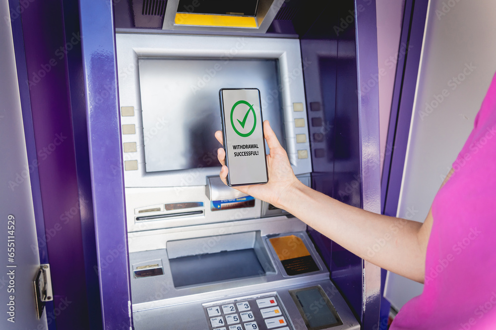 Young woman using the smart phone for cardless withdrawing the cash near the ATM Young woman using the smart phone for cardless withdrawing the cash near the ATM