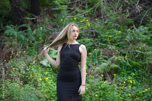 Young, beautiful blonde woman dressed in black walks through the forest in different postures and expressions. In the background ferns and yellow flowers. Concept expressions in nature.