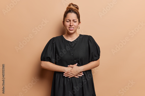 Stomachache concept. Young European woman touching belly feeling bad frowns face wears black dress gastritis disease symptom wears black dress isolated over brown background. Abdominal disease