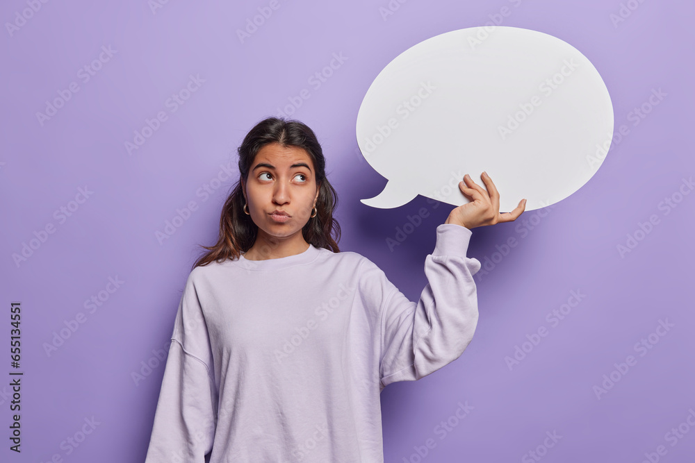 Hmm let me think what to write here. Pensive dark haired woman holds empty speech bubble concentrated aside with thoughtful expression dressed in casual sweatshirt isolated over purple background Hmm let me think what to write here. Pensive dark haired woman holds empty speech bubble concentrated aside with thoughtful expression dressed in casual sweatshirt isolated over purple background