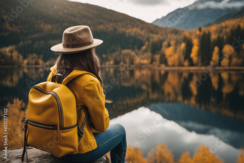 Rear view of a stylish girl, with a backpack, a hat and a yellow jacket, looking at the view of the mountains and the lake while relaxing in the autumn nature. Travel concept