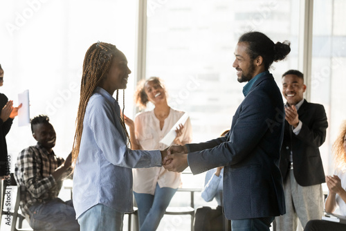 Smiling African American business partners shaking hands at meeting side view, colleagues applauding on background, executive team leader congratulating employee with promotion at briefing