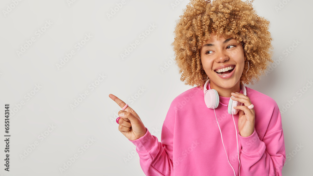 Horizontal shot of curly woman dressed in pink pullover has stereo headphones around neck for listening music enjoys spare time points at blank space for your promotion isolated over white background