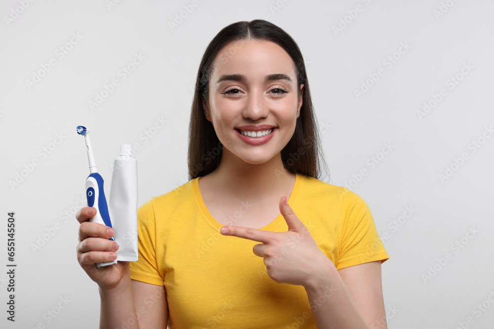 Happy young woman holding electric toothbrush and tube of toothpaste on white background Happy young woman holding electric toothbrush and tube of toothpaste on white background