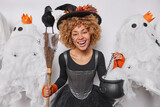Halloween party celeration. Positive curly haired young woman dressed in black dress and wizard hat has festive mood poses against spooky creatures and decoration isolated over white background