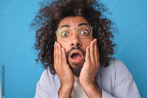 Young shocked expressive Arabian man with mental disabilities looks at screen and opens mouth suffering from panic and fear caused by bad news stands in blue studio. Close-up portrait