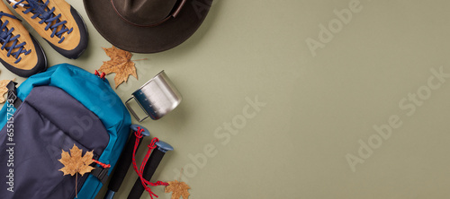 Engaging in hiking to relish the fall season. Top view flat lay of stylish hat, metal cup, trekking poles, boots, blue rucksack, fallen leaves on pastel green background with promo zone