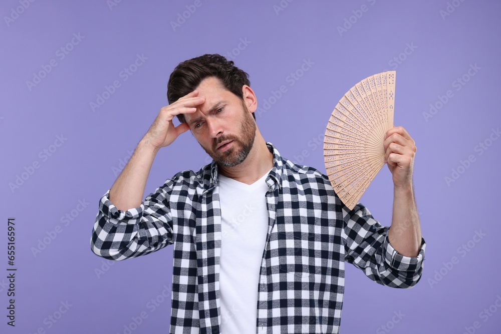 Unhappy man with hand fan suffering from heat on purple background Unhappy man with hand fan suffering from heat on purple background