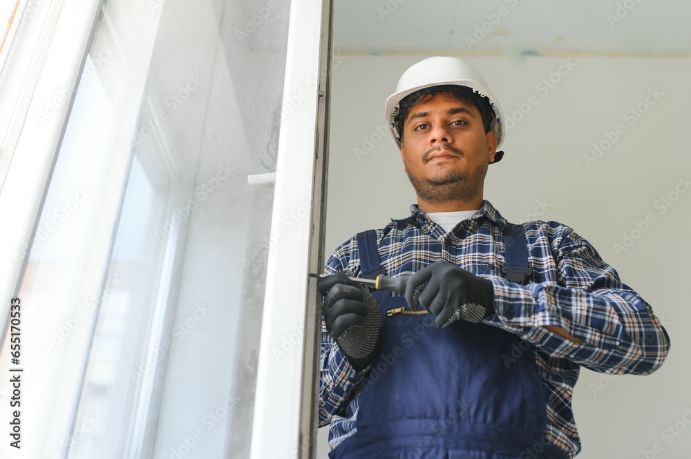 Indian Workman in overalls installing or adjusting plastic windows in the living room at home Indian Workman in overalls installing or adjusting plastic windows in the living room at home