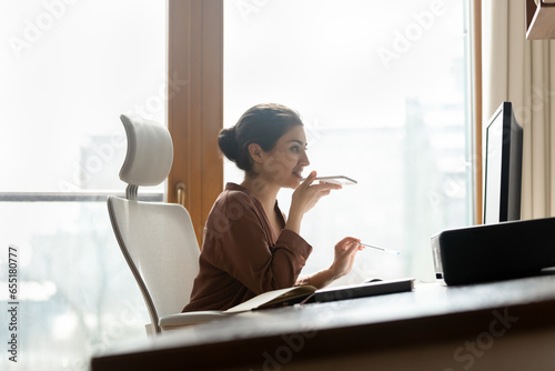 Side view smiling Indian businesswoman recording voice audio message on smartphone, sitting at workplace, looking at computer screen, chatting online by speakerphone, activating digital assistant photo