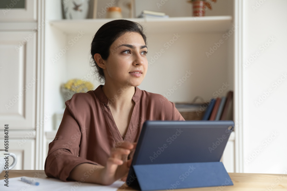 Dreamy Indian woman distracted from tablet, looking in distance, sitting at desk with modern electronic device, pensive young businesswoman or student visualizing good future, dreaming of success Dreamy Indian woman distracted from tablet, looking in distance, sitting at desk with modern electronic device, pensive young businesswoman or student visualizing good future, dreaming of success