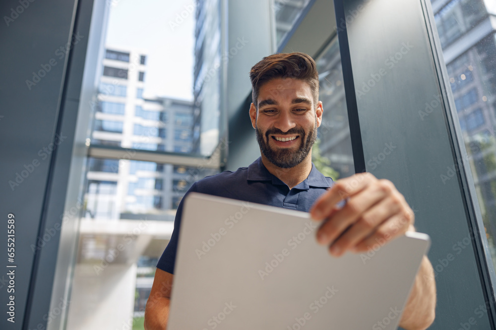 Fototapeta premium Handsome smiling freelancer working on laptop while standing in coworking near windows