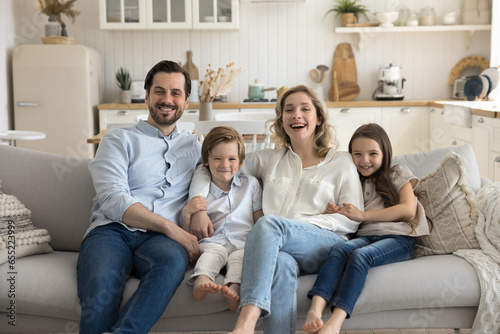 Cheerful young attractive couple with little children sit on couch in studio apartment smile, pose, look at camera, hug, show love, unity and affection. Happy homeowners family portrait, parenthood