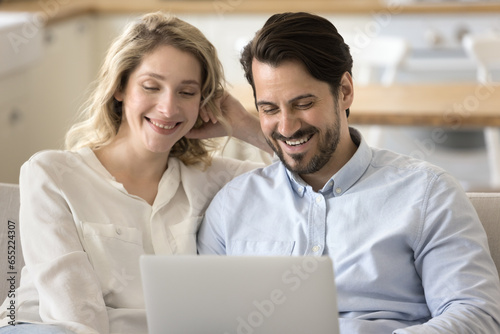 Pretty millennial 35s couple laugh while watch movie use TV streaming services, choose goods, buy travel tour, booking hotels planning joint holiday, enjoy carefree weekend time on internet use laptop
