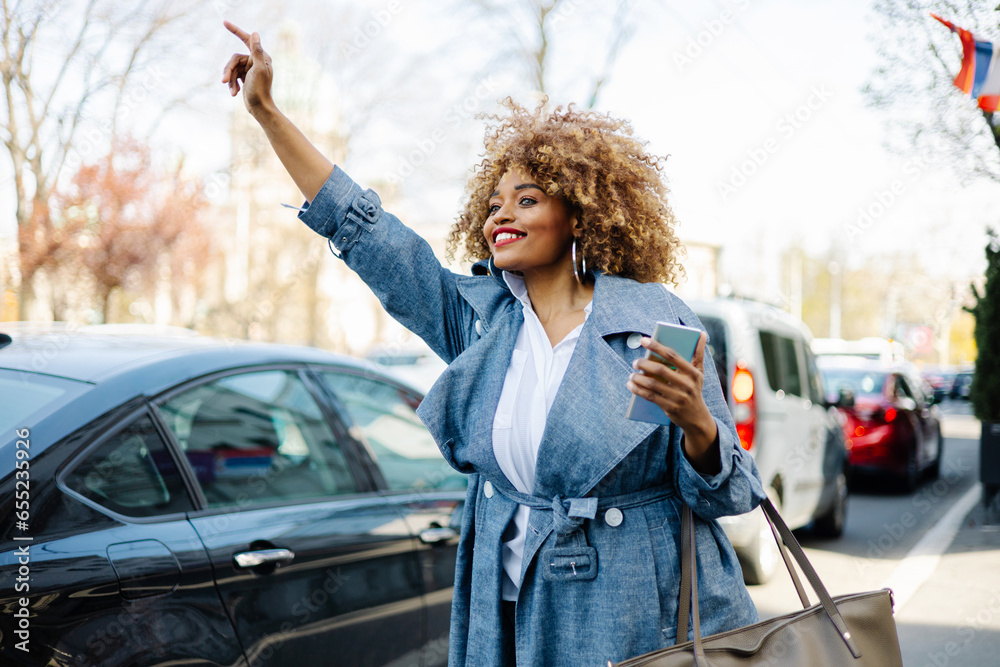Beautiful black woman walks down the street and uses smart phone for communication and calling for taxi with raised arm. She is happy and smiled. Bright sunny day. - obrazy, fototapety, plakaty 