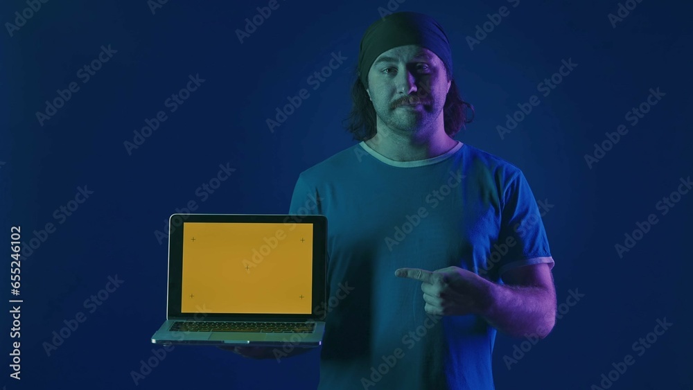 The man points to the green laptop screen in his hands. A man with a laptop in the studio on a blue background in pink and green neon light. Advertising area, workplace. The man points to the green laptop screen in his hands. A man with a laptop in the studio on a blue background in pink and green neon light. Advertising area, workplace.