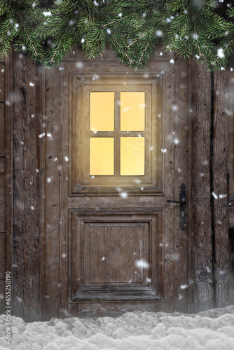 Tür mit beleuchtetem Fenster in einer verschneiten Berghütte mit Tannenzweigen