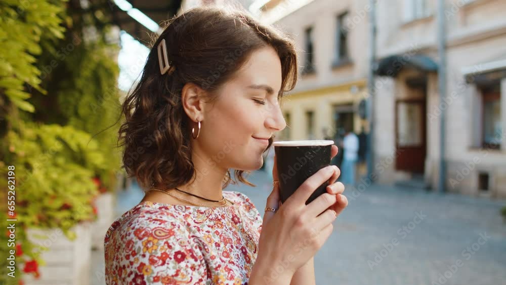 Happy Caucasian young woman enjoying morning coffee hot drink and smiling outdoors. Relaxing, taking a break. Girl walking in urban city center street, drinking coffee to go. Town lifestyles outside