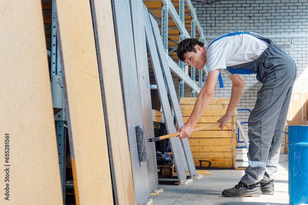 Professional industrial painter paints wooden boards with paint roller. Caucasian worker in overalls paints wooden surface indoors. Professional industrial painter paints wooden boards with paint roller. Caucasian worker in overalls paints wooden surface indoors.
